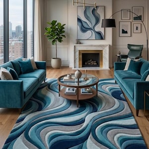 May include: A large area rug featuring a flowing, wavy pattern in shades of blue, teal, and gray. The rug is positioned in a living room between two teal velvet sofas, with a round glass-topped coffee table placed centrally on its surface.
