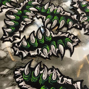 May include: Black embroidered patches featuring a claw design with sharp white claws and green accents. The patches have a detailed, textured appearance, with a black border. The claw design is repeated multiple times.