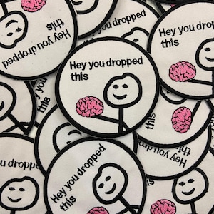 Puede incluir: Un conjunto de parches redondos blancos con bordes negros. Cada parche presenta una figura de palo sosteniendo un cerebro rosa, con el texto "Hey you dropped this" en negro. Los parches están dispersos, creando un diseño humorístico.