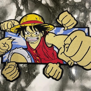 以下が含まれることがあります： 麦わら帽子と赤いベストを着た漫画のキャラクターが、青と白の背景を突き破っている刺繍パッチ。キャラクターの拳も境界線を突き破っています。パッチは黒いアウトラインと黄色、赤、白のディテールがあります。