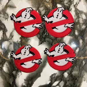 May include: Four embroidered Ghostbusters logo patches. Each patch features a red circle with a white ghost figure, crossed out with a red line. The ghost has outstretched arms and a surprised expression. The patches are displayed on a marble-effect surface.