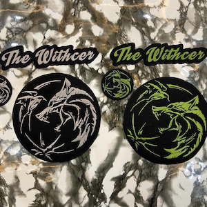 May include: Patches featuring the text "The Witcher" in black and green, along with circular patches displaying a wolf's head design. The patches are black with white or green embroidery.