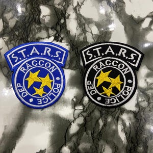 以下が含まれることがあります： ビデオゲームシリーズ、バイオハザードのS.T.A.R.S.ロゴが刺繍された2つのパッチ。1つは白と黄色のアクセントが付いた青色で、もう1つは白と黄色のアクセントが付いた黒色です。パッチには「S.T.A.R.S. Raccoon Police Dept.」の文字が書かれています。