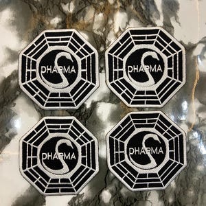 Puede incluir: Cuatro parches de la Iniciativa Dharma. Cada parche es octogonal con fondo negro y borde blanco. El centro presenta un símbolo de cisne blanco y la palabra "DHARMA". Los parches están sobre una superficie marmolada.