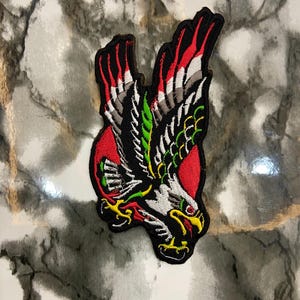 Puede incluir: Parche bordado con un águila estilizada con las alas extendidas. El parche tiene un contorno negro con detalles en rojo, blanco, verde y amarillo. El águila está sobre un fondo circular rojo.