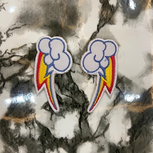 May include: Two embroidered patches featuring a cloud with a rainbow and lightning bolt design. The cloud is white with a blue outline, and the rainbow includes red, yellow, and blue. The patches are likely for clothing or accessories.