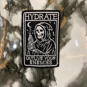 May include: Black and white embroidered patch with the text "HYDRATE" and "OUTLIVE YOUR ENEMIES". The patch depicts a Grim Reaper-like figure holding a water bottle. The background has a marble pattern.
