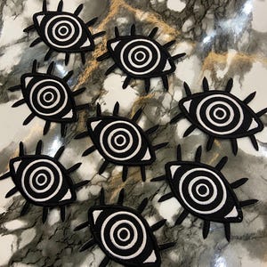 Peut inclure: Plusieurs écussons en forme d'œil noirs et blancs. Chaque écusson présente un iris en spirale blanche dans un contour noir, entouré d'épines noires. Les écussons sont disposés sur une surface marbrée.