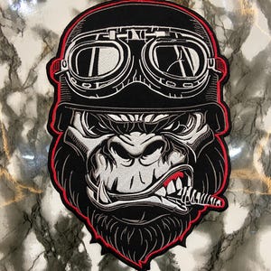 Puede incluir: Parche bordado con un gorila que lleva un casco y gafas de aviador negros. El gorila tiene una expresión feroz, un cigarro en la boca y está delineado en rojo. El parche es negro, blanco y rojo.