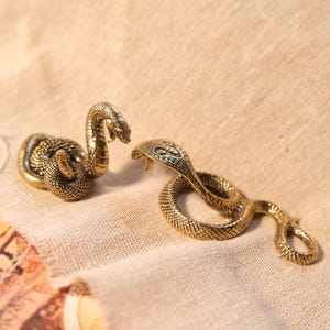 May include: Two small, metallic gold-coloured snake figurines with detailed scale textures. One snake is coiled in an upright position with its head raised, while the other snake is positioned flat in a winding, serpentine shape with its head extended.