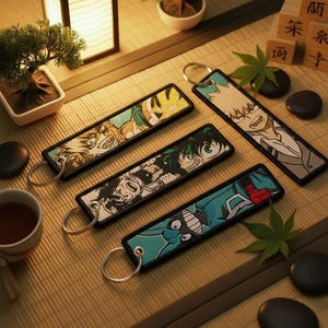 Könnte beinhalten: Vier rechteckige Schlüsselanhänger mit Anime-Charakter-Illustrationen. Jeder Schlüsselanhänger hat einen schwarzen Rand und einen silbernen Schlüsselring. Die Charaktere sind in leuchtenden Farben dargestellt, mit detaillierten Gesichtsausdrücken und dynamischen Posen. Die Schlüsselanhänger sind auf einer Bambusmatte angeordnet.