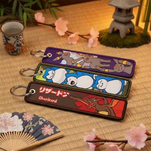 Peut inclure: Porte-clés brodés avec des personnages Pokémon. Les porte-clés sont rectangulaires avec un anneau en métal. Le porte-clés supérieur est violet avec des personnages gris et jaunes. Le porte-clés du milieu est vert avec des personnages blancs et bleus. Le porte-clés inférieur est marron avec un personnage rouge et marron et du texte japonais.