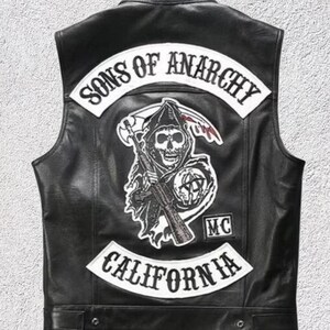 May include: A black leather vest featuring a large back patch with the text Sons of Anarchy in white. A central graphic depicts a skull in a hooded cloak holding a rifle and a scythe. A smaller patch reads MC, and a bottom patch says California.