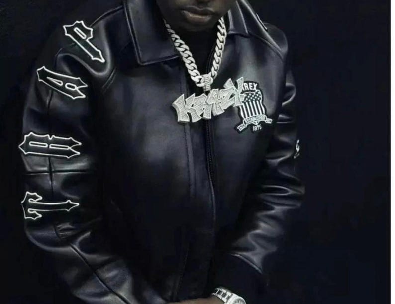 May include: Black leather jacket with white lettering on the sleeves and a patch on the chest. The jacket has a collar and a zipper closure. A silver chain necklace and a watch are also visible.