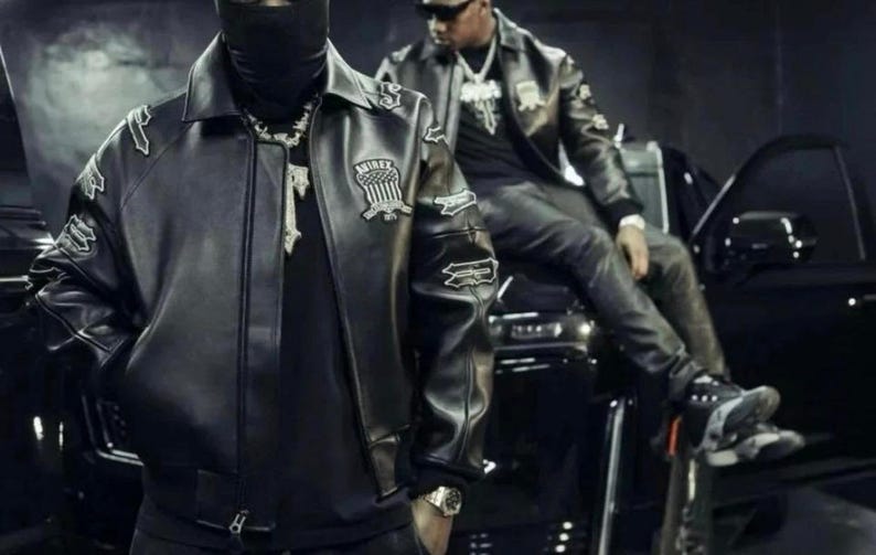 May include: Two people wearing black leather jackets with patches and a black balaclava. The jackets have a bomber style with a collar and are paired with black clothing. One person is wearing a silver watch and a silver chain.