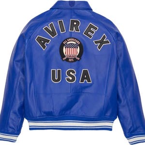K&ouml;nnte beinhalten: Eine leuchtend blaue Lederjacke mit dem Wort "AVIREX" in wei&szlig;en Buchstaben &uuml;ber den Schultern. Ein kreisf&ouml;rmiger Aufn&auml;her zeigt "ALL AMERICANS" und die US-Flagge. Das Wort "USA" ist darunter prominent platziert.