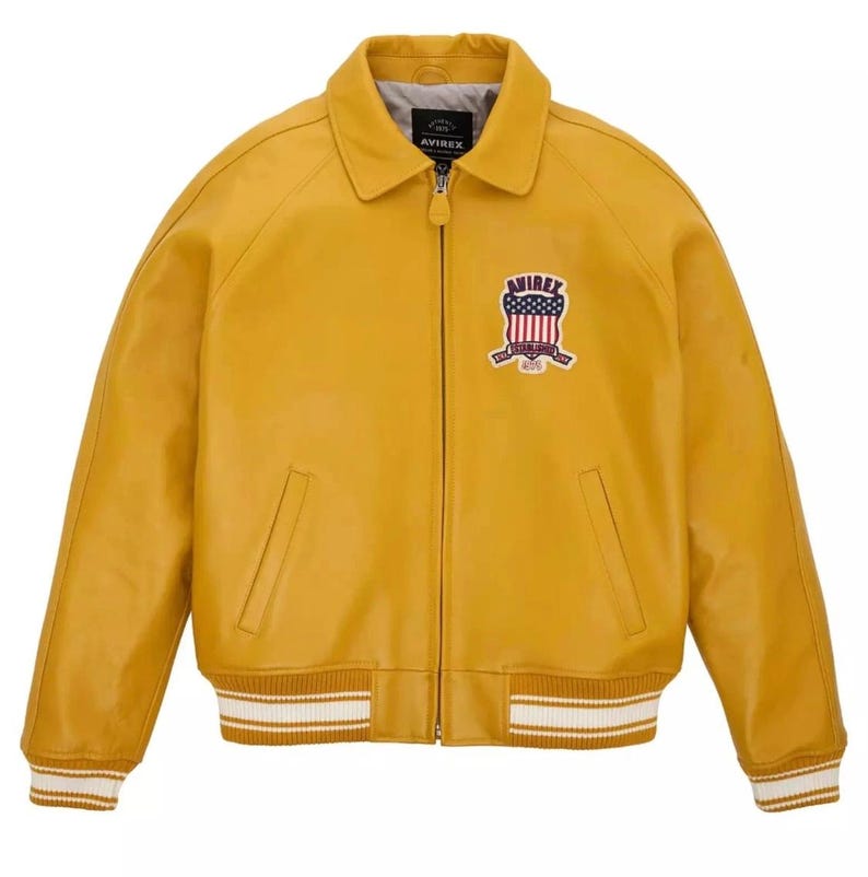 K&ouml;nnte beinhalten: Senfgelbe Lederjacke mit Rei&szlig;verschluss, Kragen und Brustaufn&auml;her. Die Jacke hat wei&szlig;-braun gestreifte B&uuml;ndchen und Saum. Der Aufn&auml;her zeigt den Text "AVIREX" und ein amerikanisches Flaggen-Design.