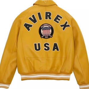 K&ouml;nnte beinhalten: Senfgelbe Lederjacke mit dem Wort "AVIREX" und "USA". Ein kreisf&ouml;rmiger Aufn&auml;her mit den Worten "ALL AMERICANS ESTABLISHED 1975" umgibt ein rot-wei&szlig;-blaues Design. Die Jacke hat einen wei&szlig;-cremefarbenen Streifenbesatz. Klassischer Bomberstil.