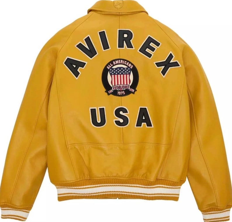 K&ouml;nnte beinhalten: Senfgelbe Lederjacke mit dem Wort "AVIREX" und "USA". Ein kreisf&ouml;rmiger Aufn&auml;her mit den Worten "ALL AMERICANS ESTABLISHED 1975" umgibt ein rot-wei&szlig;-blaues Design. Die Jacke hat einen wei&szlig;-cremefarbenen Streifenbesatz. Klassischer Bomberstil.