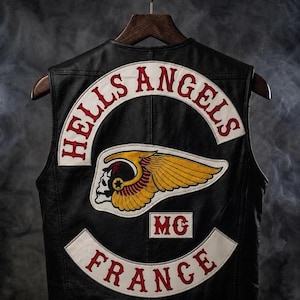 Peut inclure: Un gilet en cuir noir avec un grand écusson brodé au dos. L'écusson affiche le texte HELLS ANGELS en rouge sur une bannière blanche incurvée, un emblème de crâne ailé jaune et rouge, les lettres MC et FRANCE en rouge sur une bannière.