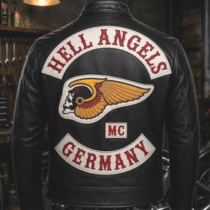 May include: A black leather jacket featuring a large embroidered patch on the back. The patch displays the text HELL ANGELS in red on a white curved background, a winged skull graphic in yellow and red, and the text GERMANY in red on a white base.