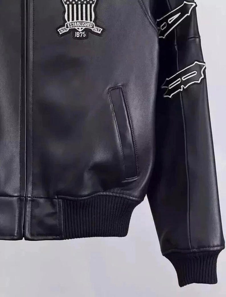 May include: Black leather jacket with a ribbed hem and cuffs. Features a patch on the chest with the text "NY ESTABLISHED 1975". White decorative stitching accents the sleeves. Includes a front pocket.