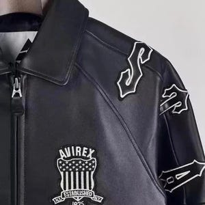 May include: Black leather jacket with a zip closure and a classic collar. Features white embroidered patches on the shoulders and sleeves. The front displays an "AVIREX" patch with "ESTABLISHED NY 1975" text.