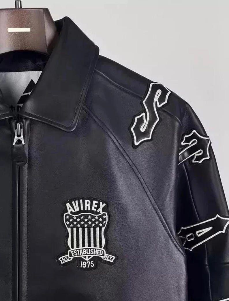 May include: Black leather jacket with a zip closure and a classic collar. Features white embroidered patches on the shoulders and sleeves. The front displays an "AVIREX" patch with "ESTABLISHED NY 1975" text.