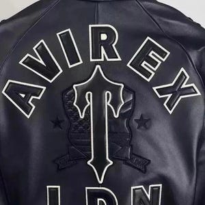 May include: Black leather jacket with white lettering and a shield emblem. The jacket features the word "AVIREX" across the top, a shield with a "T" design, and "LDN" at the bottom. Two stars flank the shield.