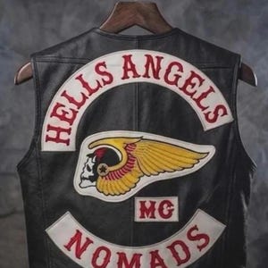Könnte beinhalten: Schwarze Lederweste mit roter Aufschrift "HELLS ANGELS" und "NOMADS". Die Weste zeigt einen geflügelten Totenkopf-Patch und die Buchstaben "MG". Sie hängt an einem Holzkleiderbügel.