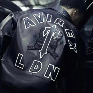May include: Black leather jacket with white lettering spelling "AVIREX LDN" on the back. A large emblem with a "T" design is centered below the word "AVIREX". The jacket has a classic bomber style.