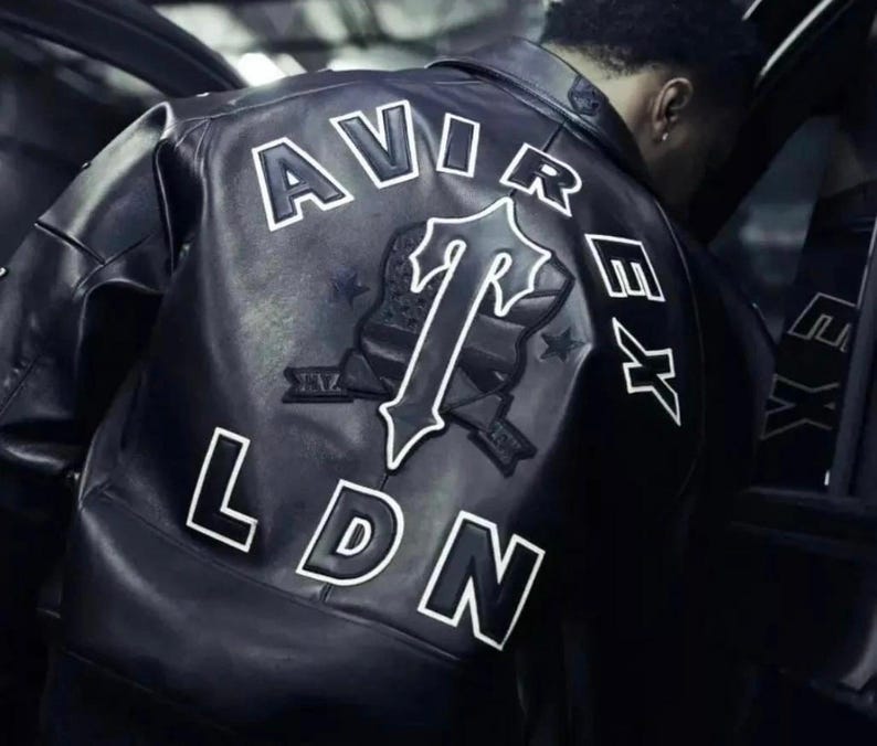 May include: Black leather jacket with white lettering spelling "AVIREX LDN" on the back. A large emblem with a "T" design is centered below the word "AVIREX". The jacket has a classic bomber style.