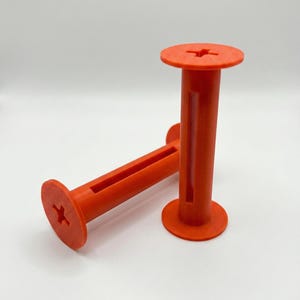 Puede incluir: Dos carretes naranjas impresos en 3D. Cada carrete tiene una base y una parte superior circulares, con un cuerpo cilíndrico y una ranura rectangular. La parte superior e inferior presentan un diseño en forma de cruz. Están diseñados para sujetar algo.