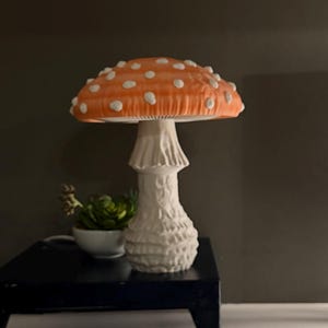 May include: A mushroom-shaped lamp featuring a vibrant orange cap with raised white spots and a textured white stalk. The lamp is positioned on a black stand next to a small potted succulent, providing a distinct light source in the room.