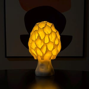Puede incluir: Una l&aacute;mpara de mesa con forma de seta que presenta una c&uacute;pula texturizada tipo panal que brilla con una luz amarilla c&aacute;lida. La base es lisa y blanca, sosteniendo la parte superior iluminada que proyecta un suave resplandor dorado.
