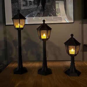May include: Three black street lamp-style lanterns of varying heights, each featuring a hexagonal base, a vertical post, and a lantern head with a diamond-patterned mesh enclosure. Each lantern contains a warm, glowing yellow light inside.