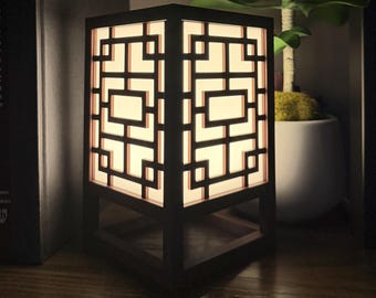 May include: A square table lamp with a dark brown wooden frame featuring a geometric lattice pattern. The lamp emits a soft, warm glow from behind a translucent white panel, supported by a sturdy open base structure on a flat surface.