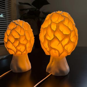 Puede incluir: Dos l&aacute;mparas con forma de hongo con bases blancas y sombreros de color naranja brillante y texturizado. Los sombreros presentan una estructura de superficie picada similar a un panal que emite una luz c&aacute;lida. Cada l&aacute;mpara tiene un cable blanco.