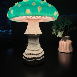 May include: A whimsical mushroom-shaped lamp with a teal cap and a white stem. The cap features yellow-orange spots and emits a soft glow. The lamp is on a dark surface, with a small vase of greenery in the background.