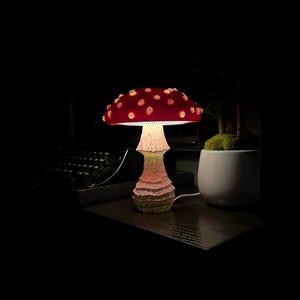 May include: A mushroom-shaped table lamp featuring a vibrant red cap with raised white spots and a textured, light-colored stalk. The lamp emits a soft glow from beneath the cap, illuminating the stem and the surrounding area in a dark environment.