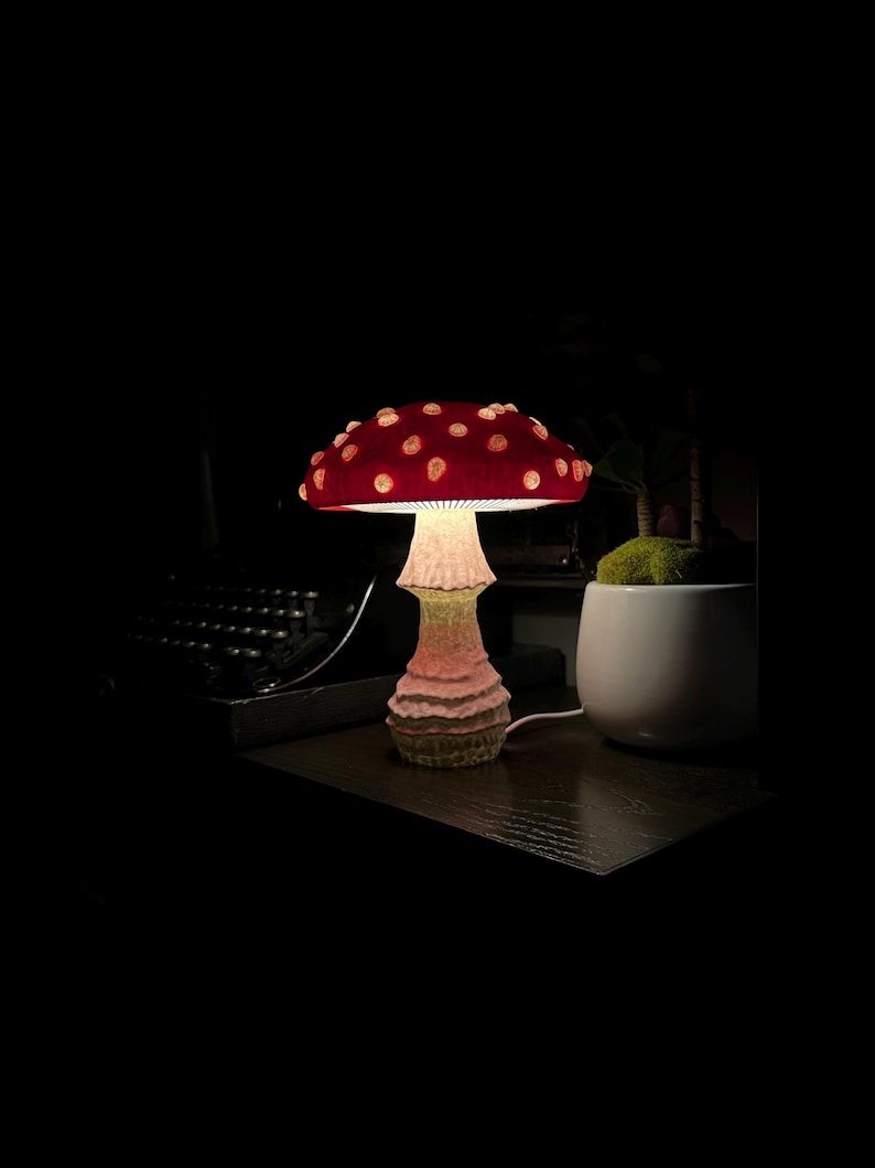 May include: A mushroom-shaped table lamp featuring a vibrant red cap with raised white spots and a textured, light-colored stalk. The lamp emits a soft glow from beneath the cap, illuminating the stem and the surrounding area in a dark environment.