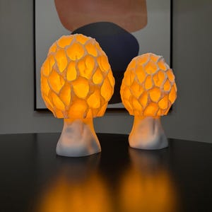 Puede incluir: Dos l&aacute;mparas con forma de seta con sombreretes blancos texturizados de patr&oacute;n de panal que emiten una luz naranja c&aacute;lida y brillante. Las l&aacute;mparas tienen bases blancas lisas y s&oacute;lidas, y se mantienen erguidas proyectando un suave resplandor &aacute;mbar.