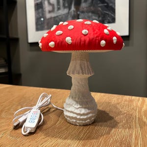 May include: A mushroom-shaped lamp with a bright red cap featuring raised white spots and a textured, off-white stalk. The lamp includes a white USB power cord with a small control panel featuring four buttons for adjusting light settings.