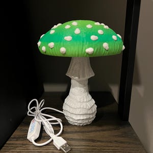 May include: A mushroom-shaped lamp featuring a vibrant green cap with white spots and a textured white stem. The lamp includes a white power cord with a USB connector and an integrated control switch for adjusting the light settings.
