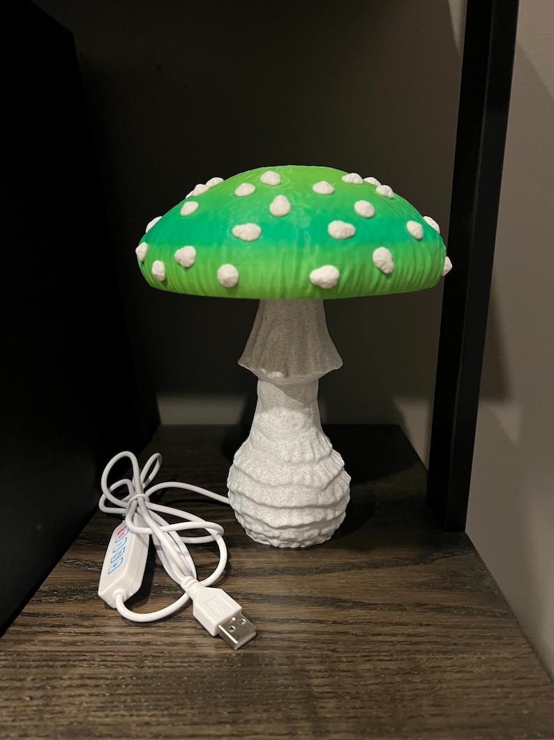 May include: A mushroom-shaped lamp featuring a vibrant green cap with white spots and a textured white stem. The lamp includes a white power cord with a USB connector and an integrated control switch for adjusting the light settings.