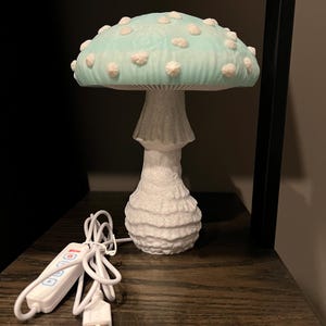 May include: A mushroom-shaped lamp featuring a light blue cap with raised white spots and a textured white stem. The lamp includes a white power cord with an integrated control panel and a USB connector for operation, providing a functional light source.