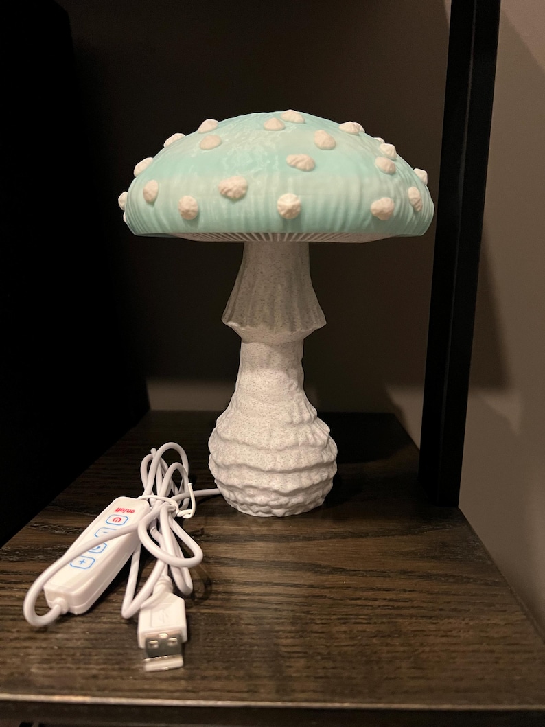 May include: A mushroom-shaped lamp featuring a light blue cap with raised white spots and a textured white stem. The lamp includes a white power cord with an integrated control panel and a USB connector for operation, providing a functional light source.