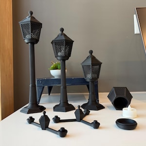 May include: Three black matte street lamp-style candle holders of varying heights, each featuring a hexagonal lantern top with a grid pattern. Included are a separate hexagonal base, a small white tea light, and a black cross-shaped connector piece.