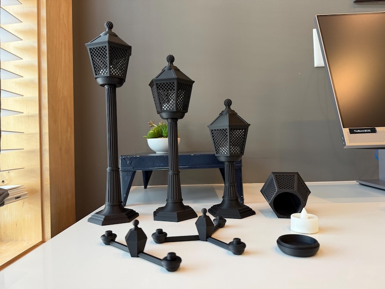 May include: Three black matte street lamp-style candle holders of varying heights, each featuring a hexagonal lantern top with a grid pattern. Included are a separate hexagonal base, a small white tea light, and a black cross-shaped connector piece.