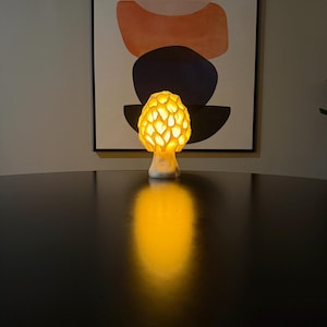 Puede incluir: Una l&aacute;mpara con forma de seta con una tapa texturizada similar a un panal que emite un c&aacute;lido resplandor amarillo dorado. La base es lisa y blanca, y la luz proyecta un reflejo brillante y alargado sobre la superficie oscura debajo.